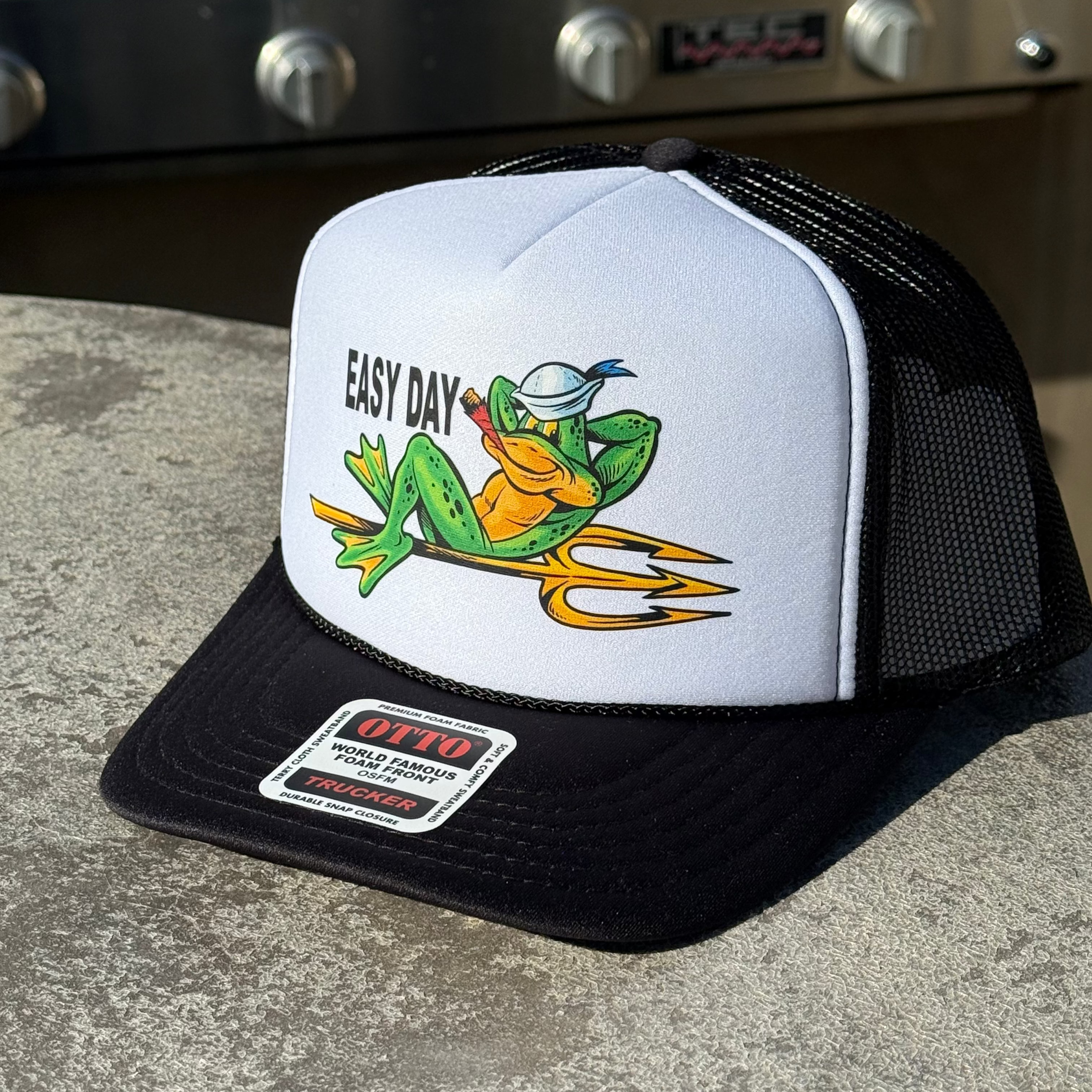 White and black cap with a frog graphic and 'Easy Day' text on a concrete surface.