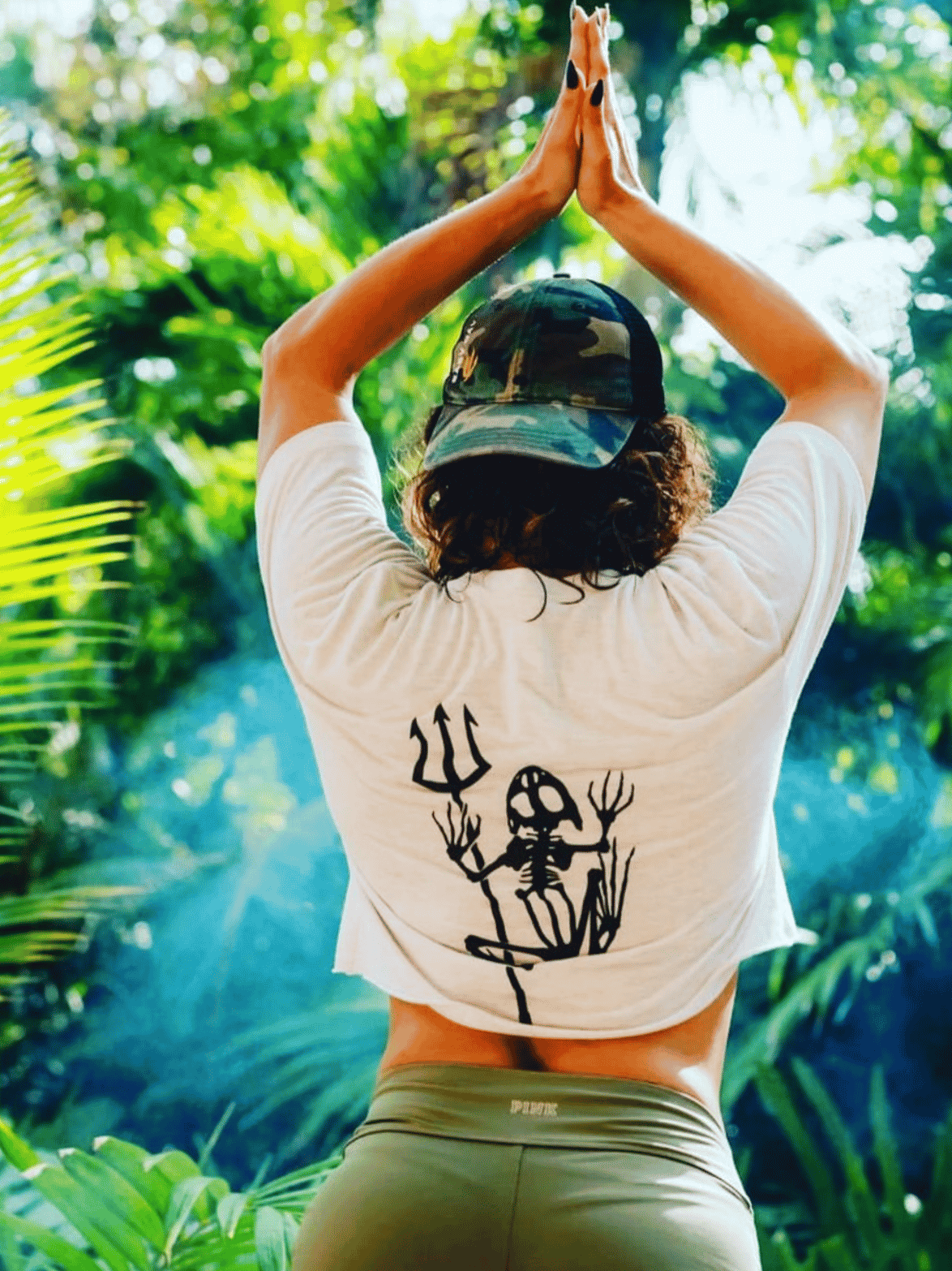 Person in a white shirt with a black design and green shorts standing in a yoga pose with hands above head in a lush green outdoor setting.