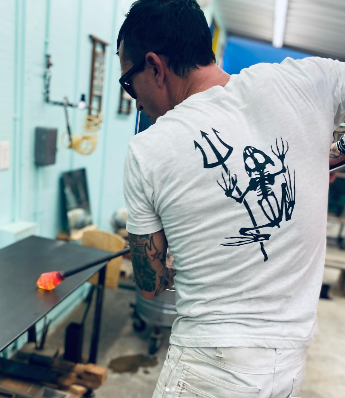 Person wearing a white t-shirt with a black easy day bonefrog design on the back, standing in a workshop.
