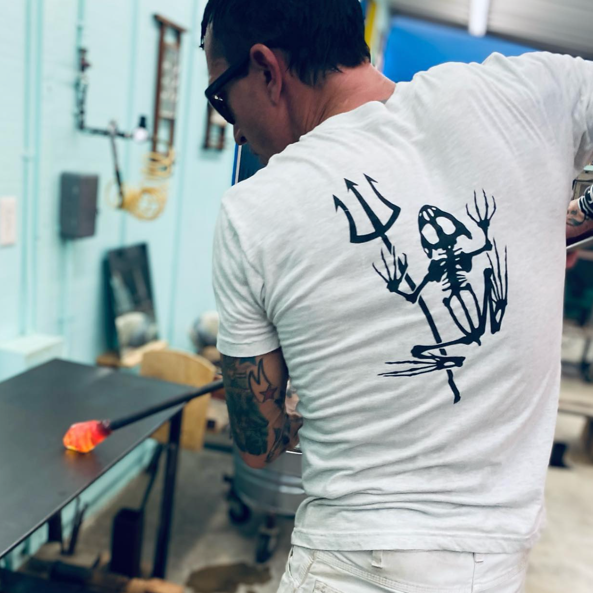 Person wearing a white t-shirt with a black easy day bonefrog design on the back, standing in a workshop.