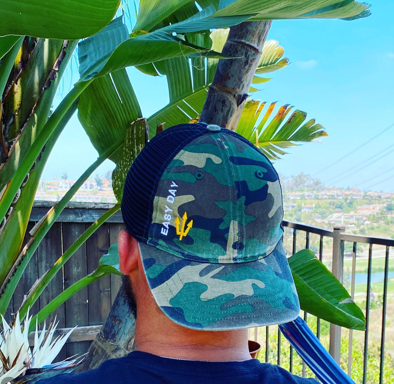 Navy SEAL Jason Higgins wearing a camouflage cap with 'Easy Day' branding, standing on a balcony with greenery and a cityscape in the background.